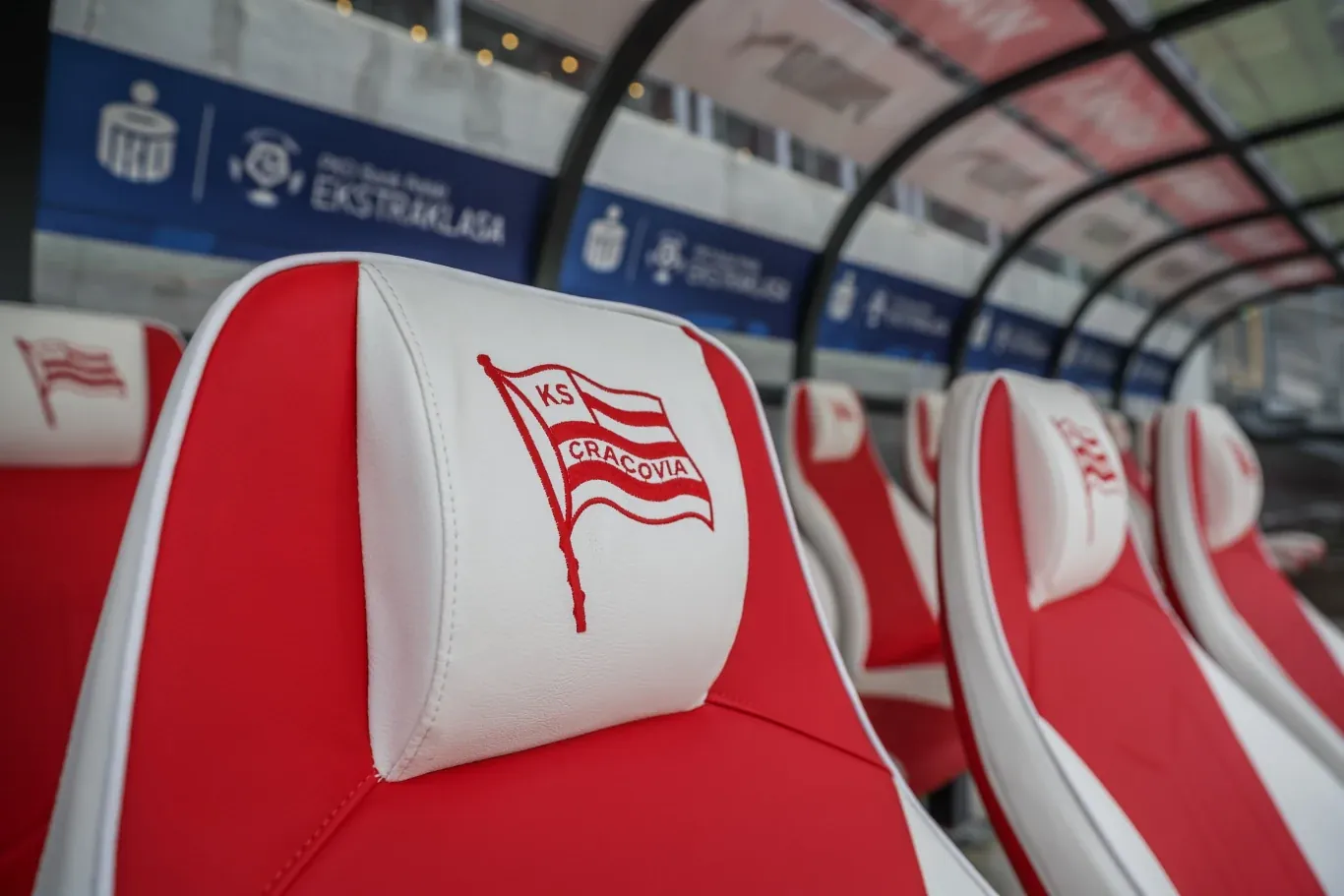 Rząd czerwono-białych krzeseł z herbem KS Cracovia w strefie technicznej stadionu piłkarskiego.