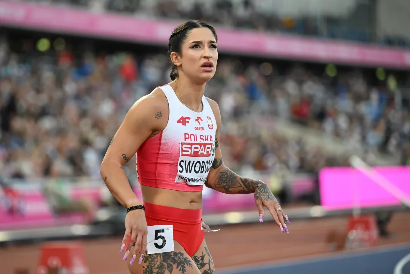 Kobieta w stroju sportowym z numerem startowym stoi na bieżni lekkoatletycznej, w tle widoczna publiczność stadionu.