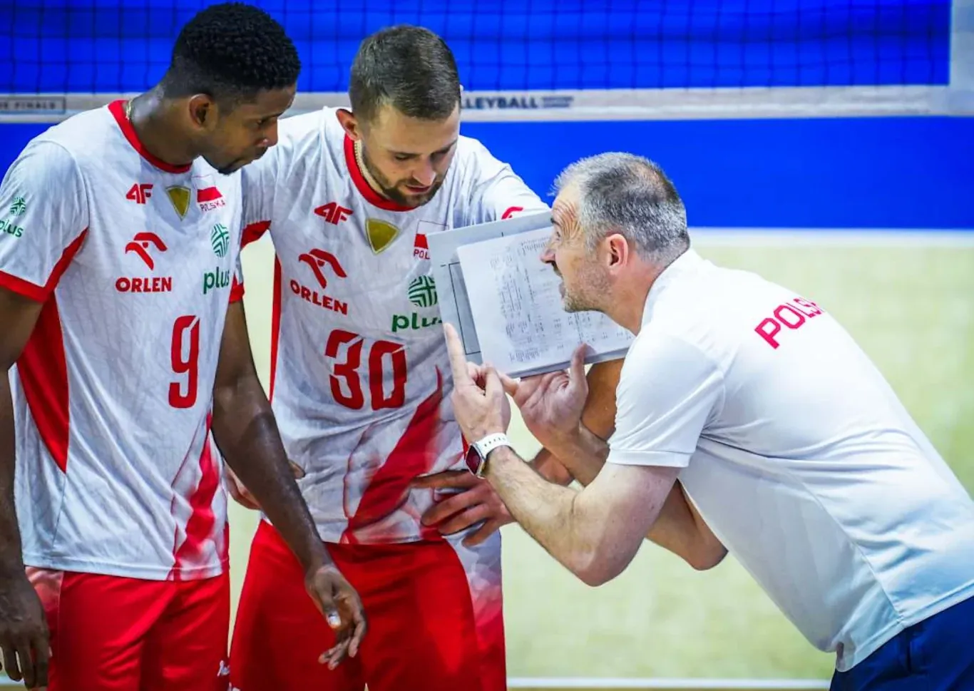 Trener drużyny siatkówki przekazuje taktyczne wskazówki dwóm zawodnikom ubranym w biało-czerwone stroje, trzymając w ręku kartkę z rozrysowaną strategią.