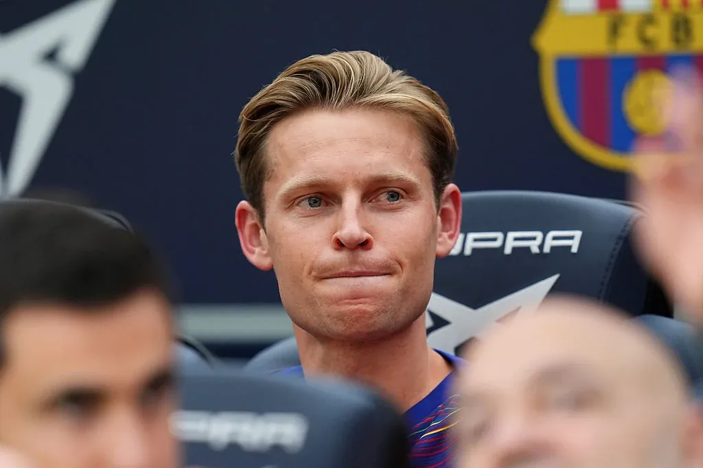 Młody mężczyzna z blond włosami siedzi na sportowym stadionie, ubrany w koszulkę w barwach klubu FC Barcelona, otoczony innymi osobami na trybunie rezerwowych.
