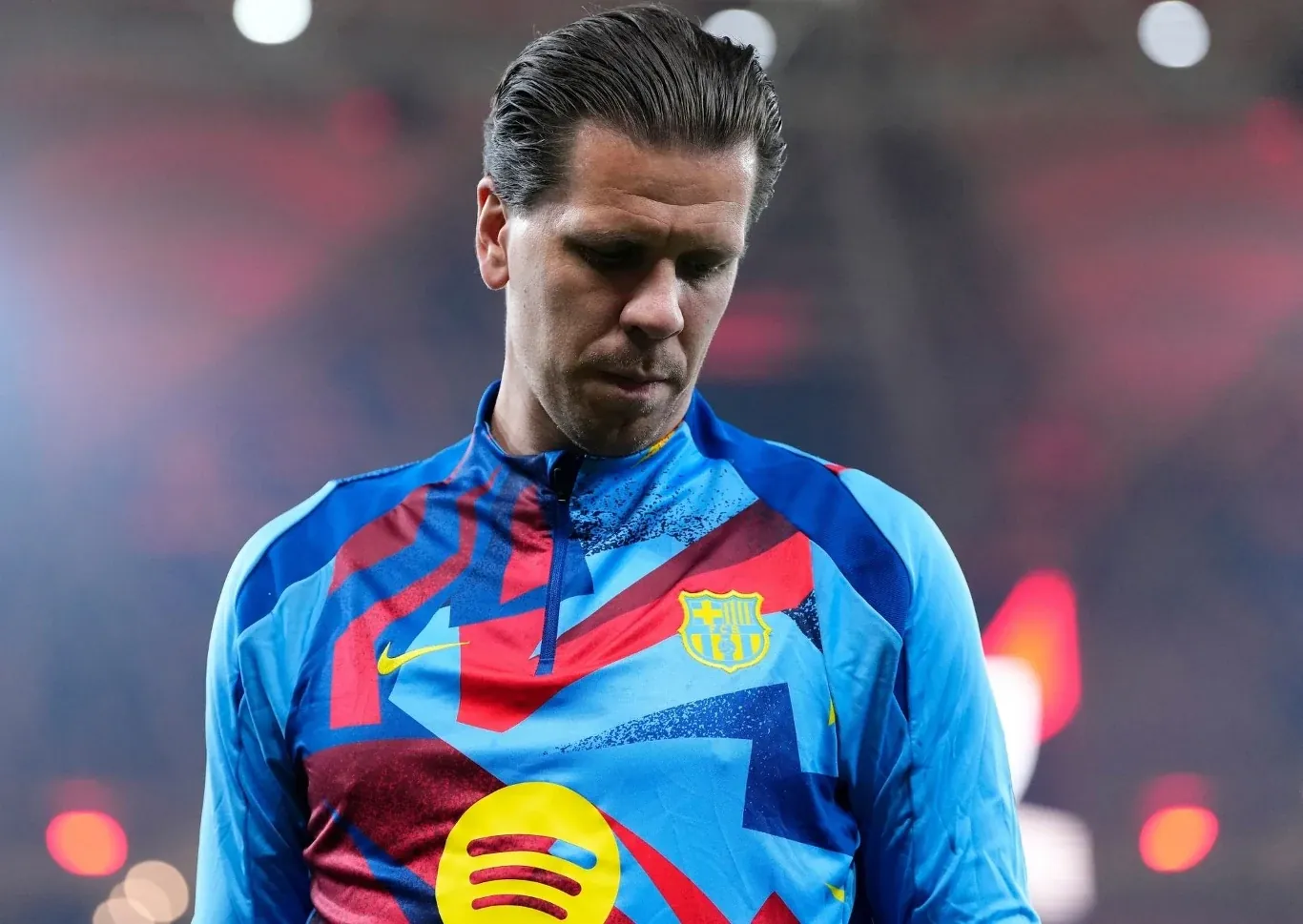 Piłkarz w kolorowym stroju sportowym FC Barcelony patrzący w dół, światła stadionu rozmyte w tle.