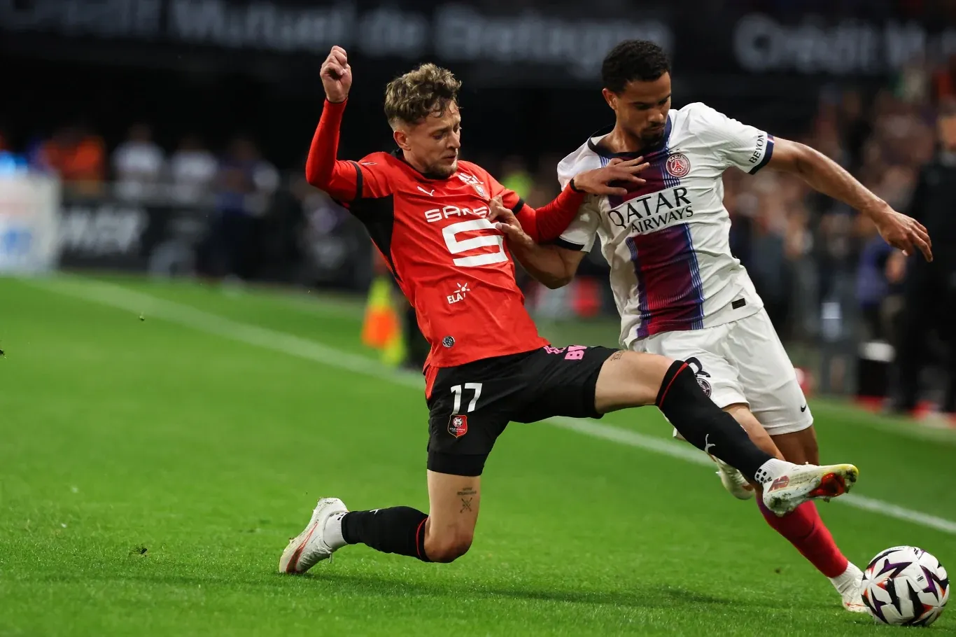 Sebastian Szymański w czerwono-czarnym stroju wykonuje wślizg przy zawodniku w białych barwach na murawie.