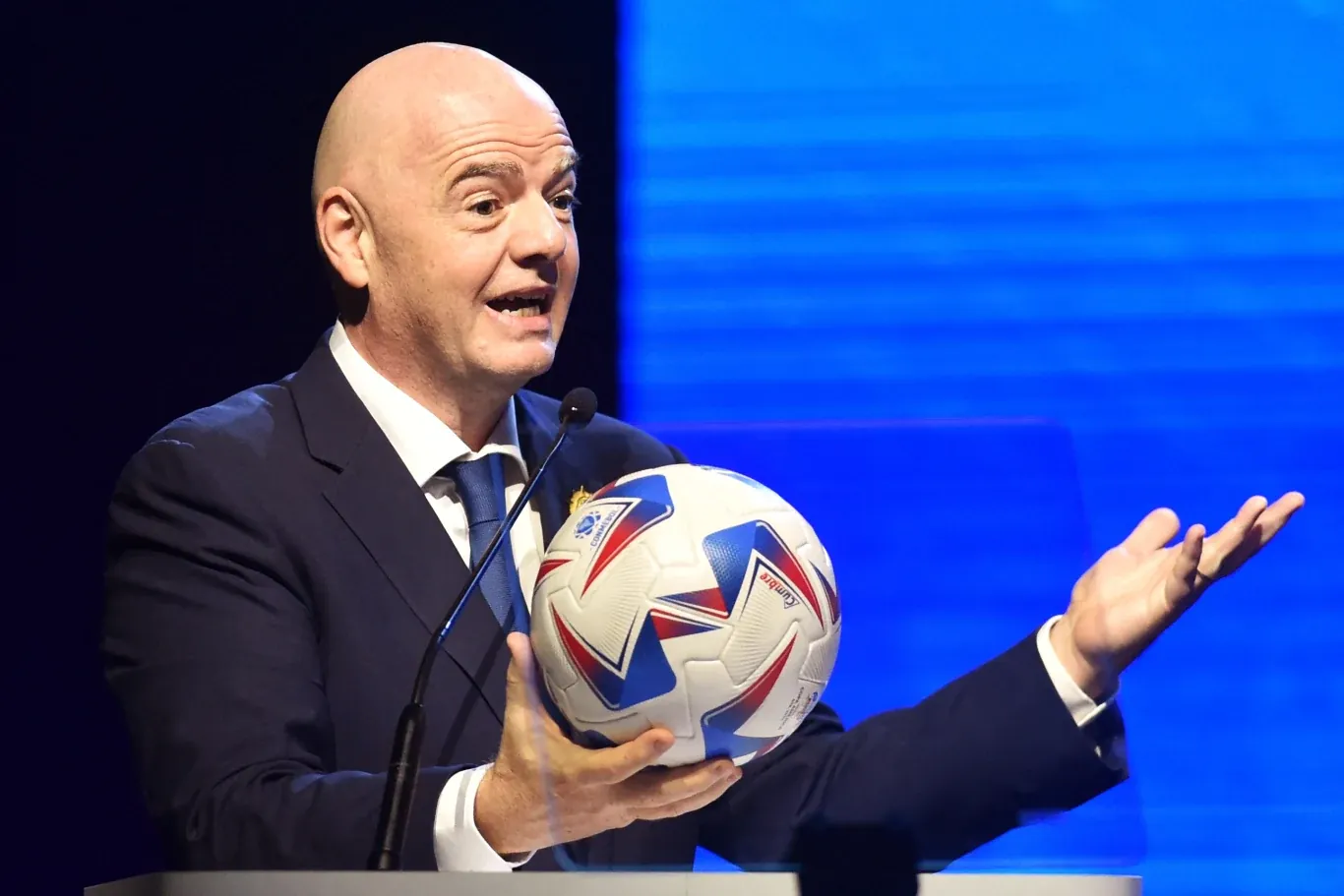 Gianni Infantino, szef FIFA, przemawia do mikrofonu, trzymając piłkę nożną na tle niebieskiego światła