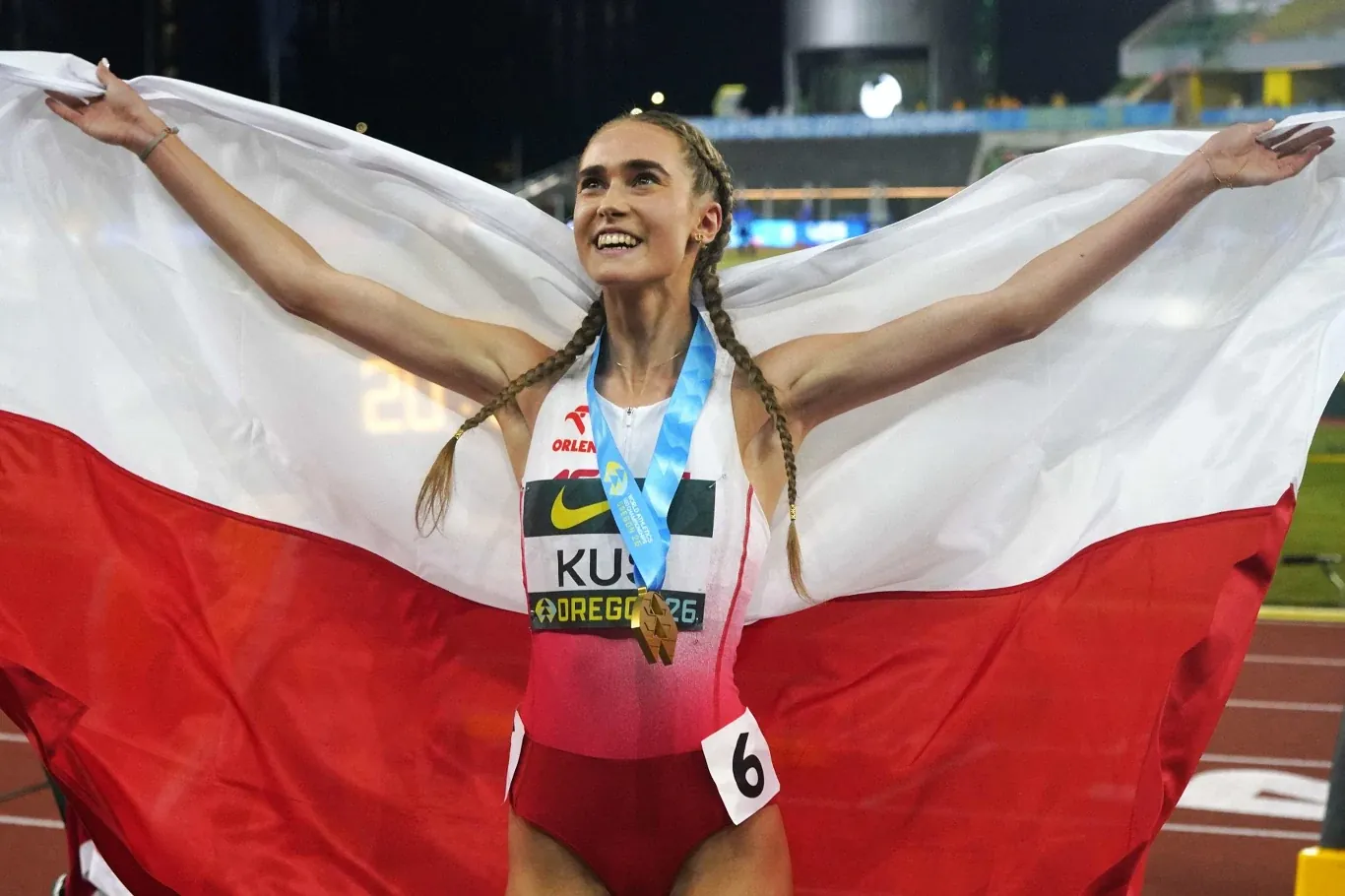 Lekkoatletka w biało-czerwonym stroju z medalem na szyi trzyma rozpostartą polską flagę i uśmiecha się szeroko po zakończonym biegu na stadionie lekkoatletycznym.
