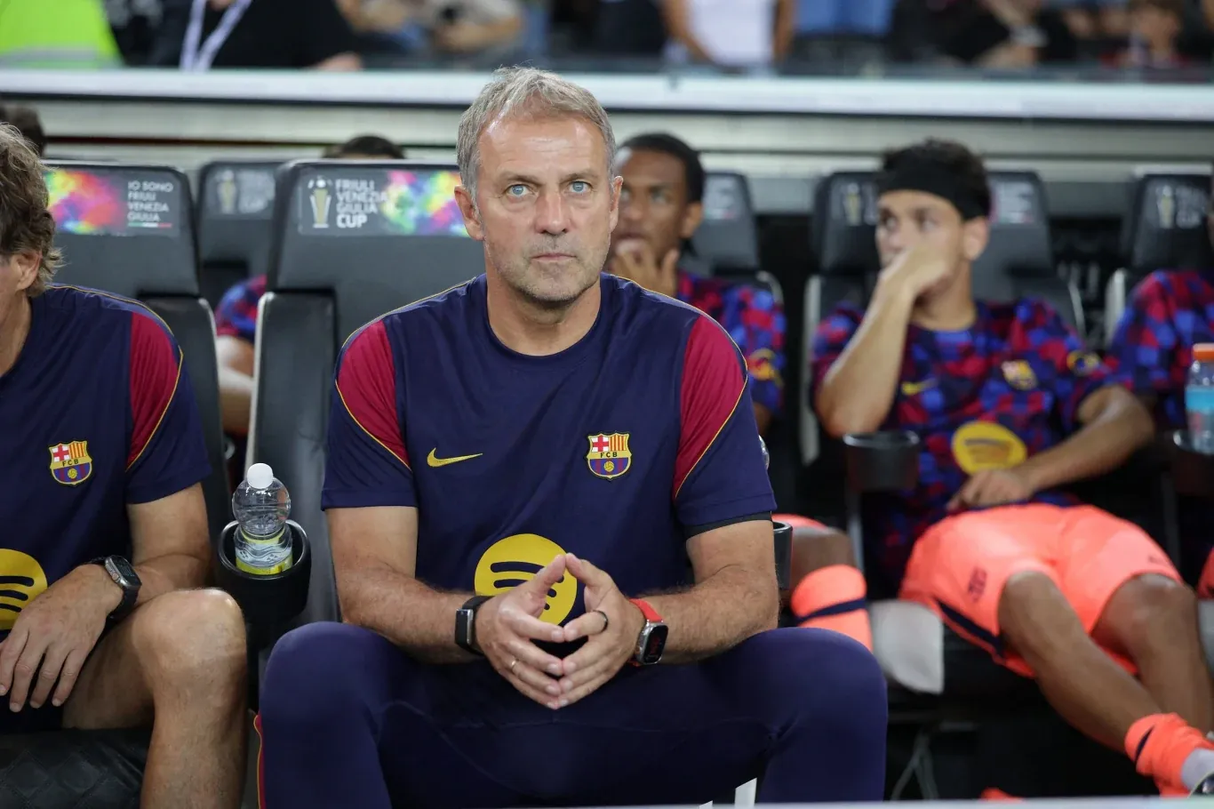 Trener drużyny FC Barcelona siedzi na ławce rezerwowych, ubrany w klubowy strój treningowy, skoncentrowany na wydarzeniach na boisku, wokół niego pozostali członkowie zespołu rezerwowych.