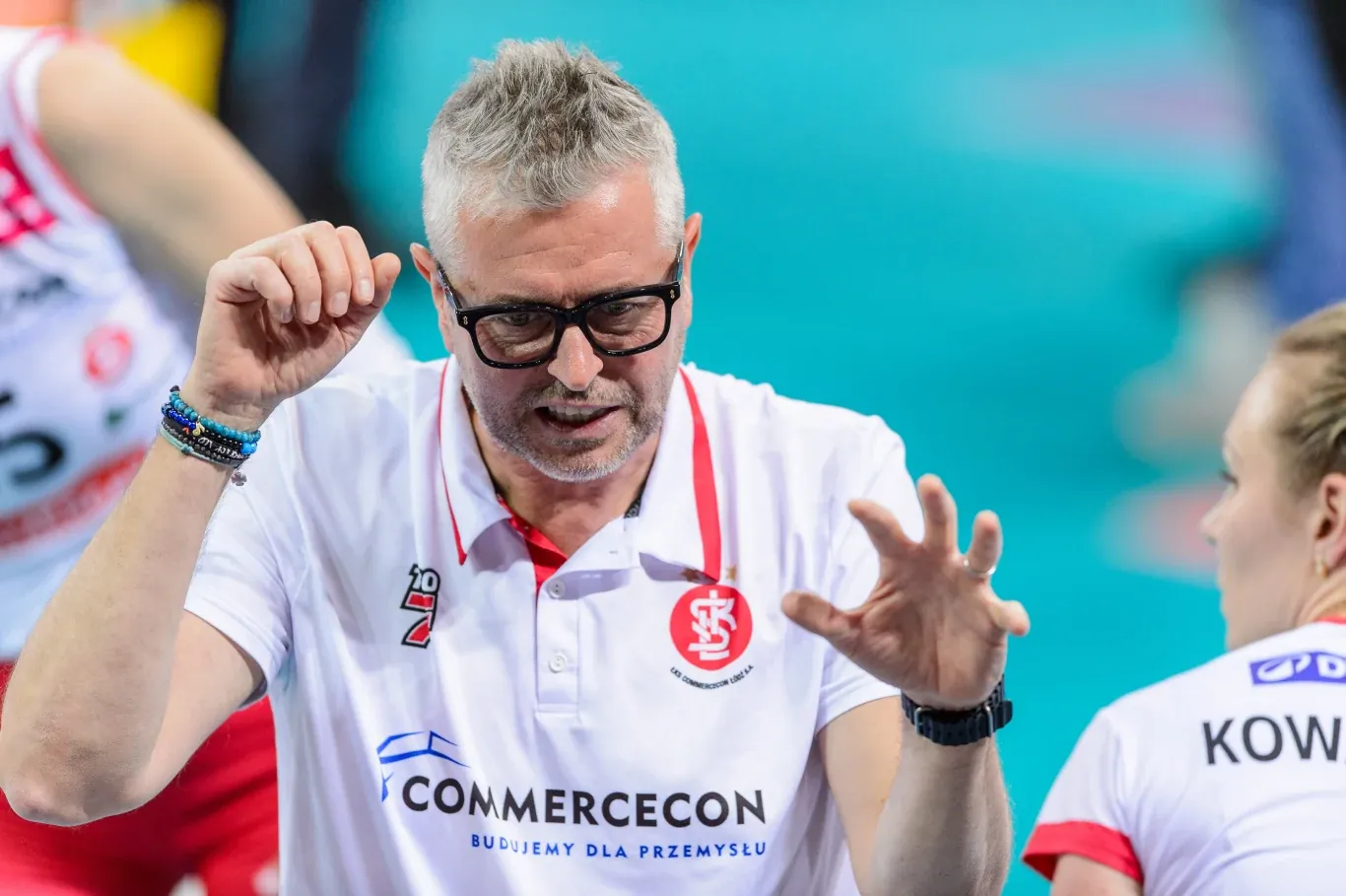 Alessandro Chiappini w jasnej koszulce ŁKS, w okularach, gestykuluje obok siatkarki podczas meczu.