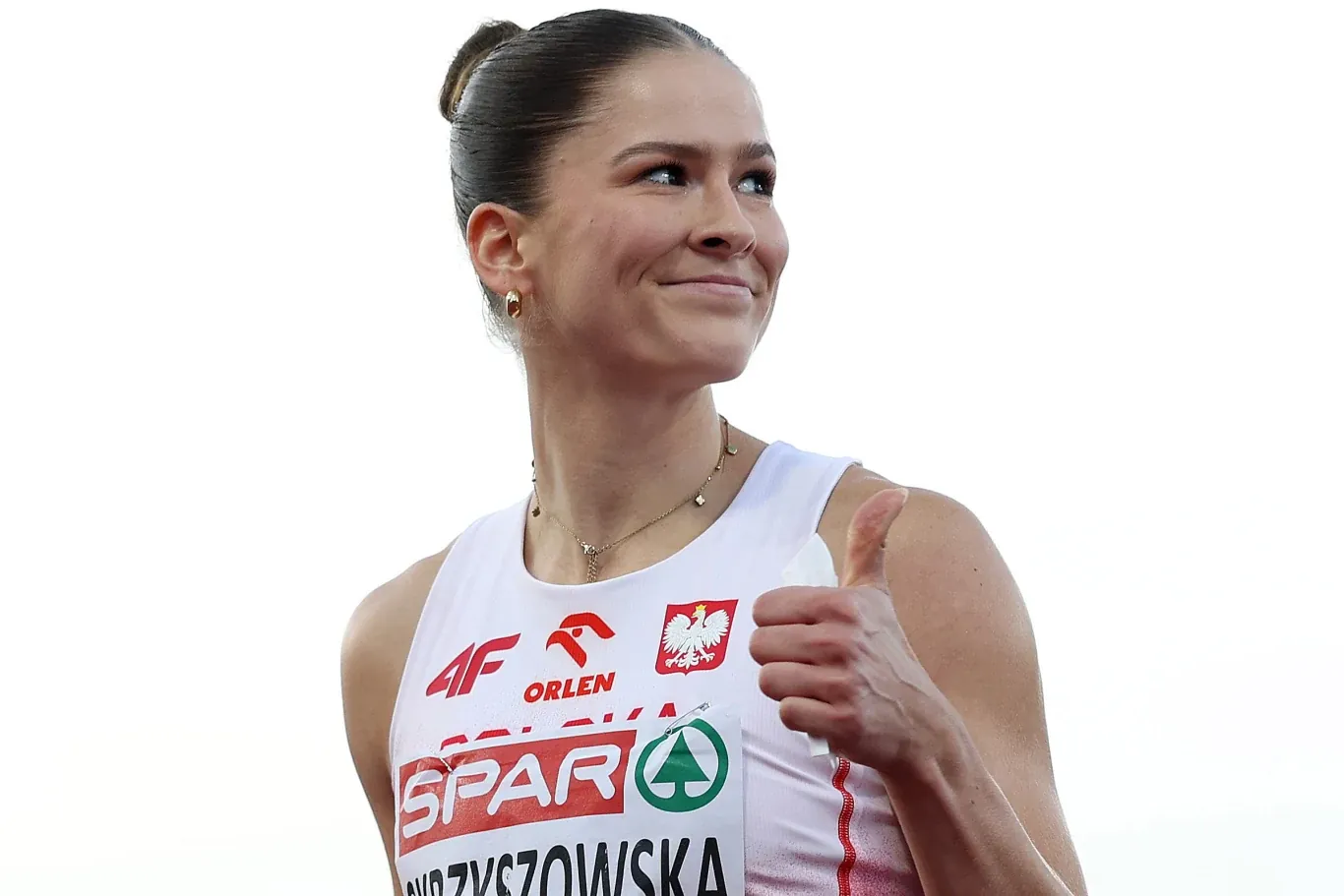 Lekkoatletka w biało-czerwonym stroju z polskim godłem oraz logotypami sponsorów, unosi kciuk do góry i uśmiecha się, emanując pozytywną energią.