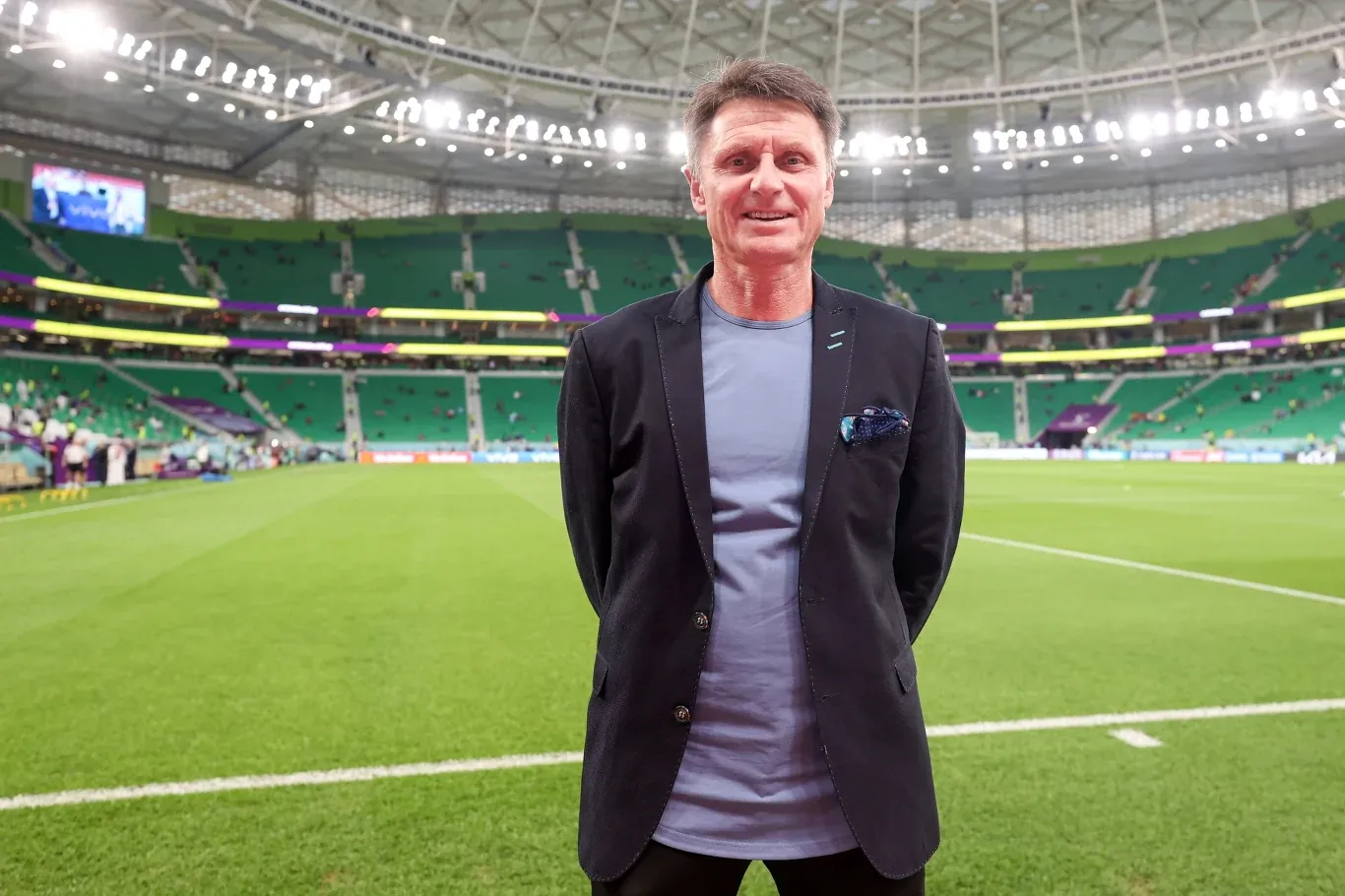 Uśmiechnięty mężczyzna w jasnej koszulce i ciemnej marynarce stoi na murawie nowoczesnego stadionu piłkarskiego z zielonymi trybunami i zadaszeniem.