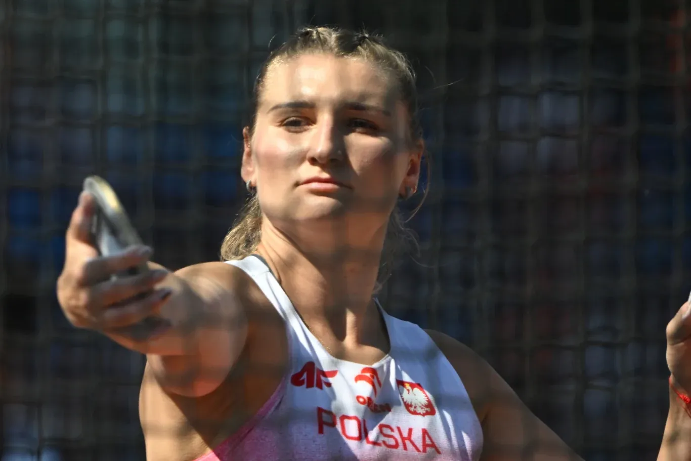 Lekkoatletka w polskim stroju sportowym skupiona przed rzutem dyskiem, widoczna zza siatki ochronnej stadionu.