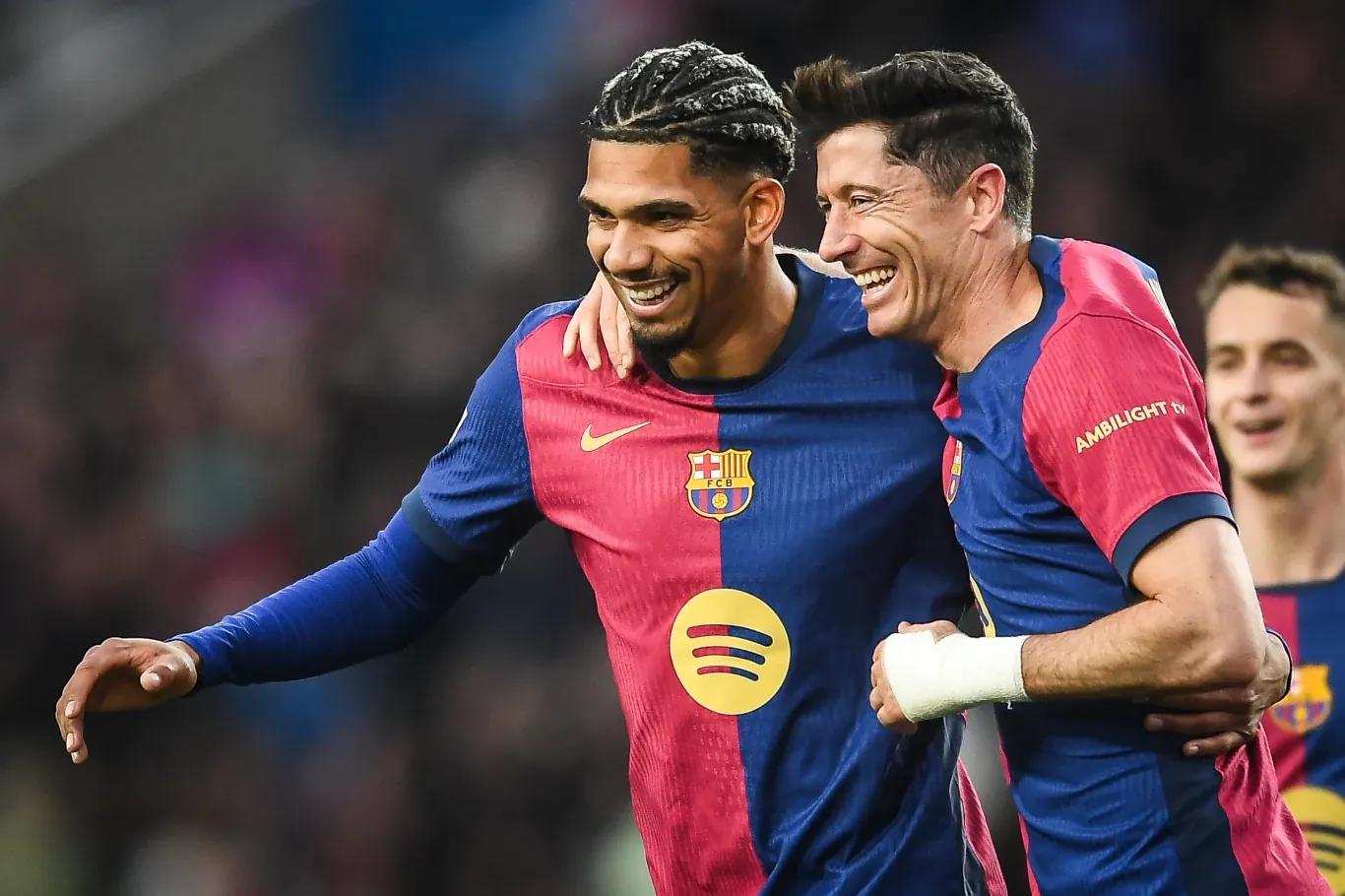 Dwóch piłkarzy FC Barcelony w charakterystycznych granatowo-bordowych koszulkach obejmuje się na boisku, uśmiechając się po udanej akcji, w tle rozmyte sylwetki innych osób.