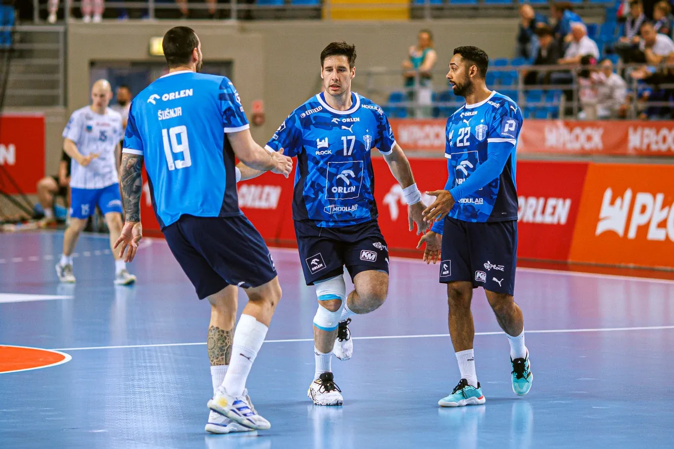 Trzech szczypiornistów w niebieskich koszulkach drużyny ORLEN Wisła Płock celebruje akcję na parkiecie hali sportowej, skupiając się na wspólnym geście przybicia piątki, wokół nich widoczni są inni zawodnicy i trybuny z kibicami.