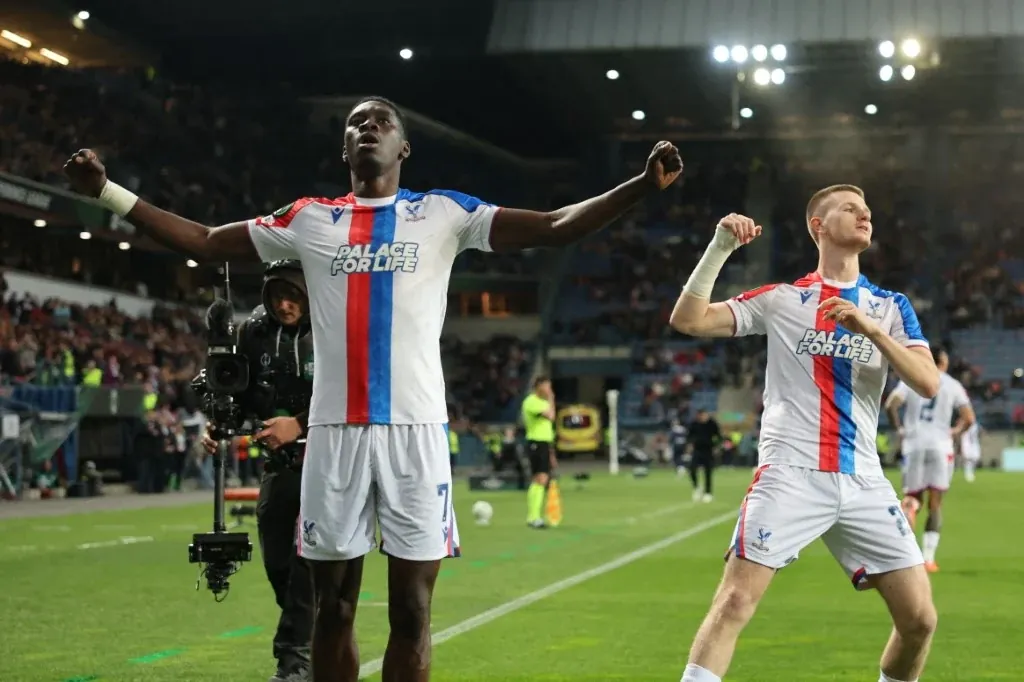 Dwóch piłkarzy w biało-niebiesko-czerwonych strojach Crystal Palace z uniesionymi rękami celebruje na murawie stadionu po zakończonej akcji, w tle widoczny tłum kibiców i pracownicy techniczni przy linii bocznej.