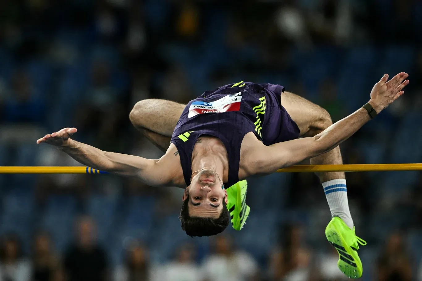 Lekkoatleta w trakcie skoku wzwyż na stadionie, z ciałem wygiętym łukiem nad poprzeczką i skupionym wyrazem twarzy, w charakterystycznych sportowych butach i stroju startowym.