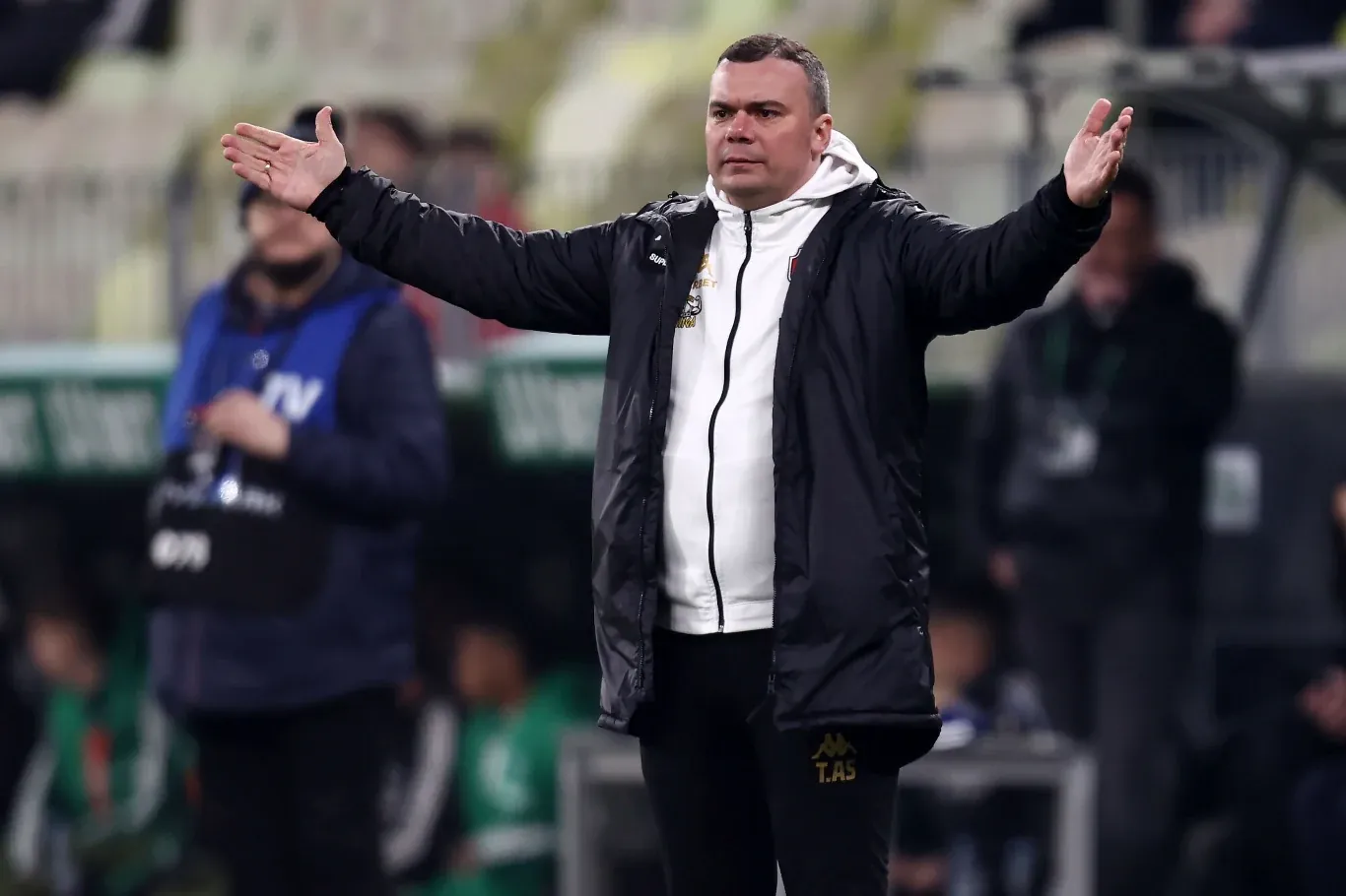 Adrian Siemieniec w czarnej kurtce i białej bluzie, z rozłożonymi rękami podczas meczu piłkarskiego